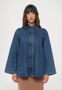 Casaquinho de ganga com colarinho estruturado, frente com fecho de botões e mangas largas. Cor azul-escuro com um acabamento ligeiramente desbotado.