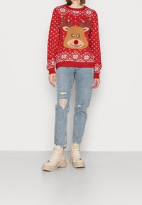 Pull rouge tricoté avec un motif de renne, un motif de flocons de neige et des boutons dorés. Associé à un jean bleu clair déchiré et des bottes beiges à lacets.