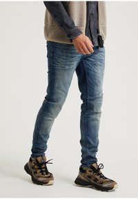 Haaleat siniset farkut kuluneilla yksityiskohdilla, yhdistetty ruskeisiin ja mustiin urheilutennareihin sekä beige fleece-liivi tumman paidan päällä.