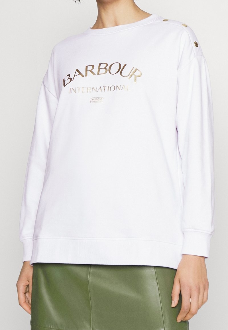 Bílá mikina s dlouhým rukávem a zlatým logem "Barbour International", s knoflíkovými detaily na rameni, nošená s olivově zelenou koženou sukní.