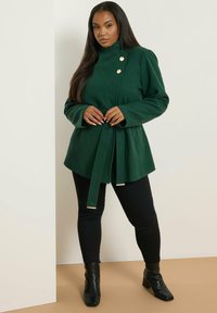 Vihreä villasekoitteinen takki, jossa on korkea kaulus, kaksoisnapitus ja vyötäröllä vyö. Yhdistetty mustiin housuihin ja nilkkureihin.