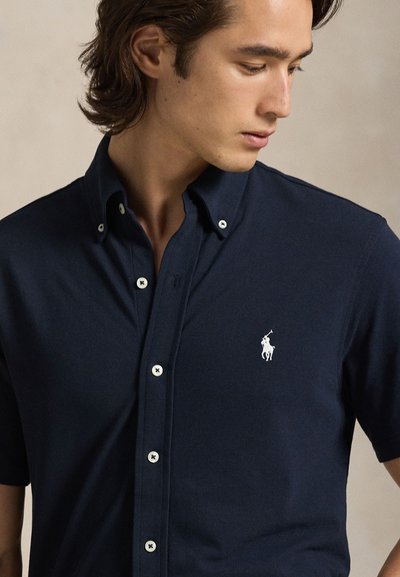 Μπλε ναυτικό polo με κοντό μανίκι από υφασμα με υφή, που διαθέτει μυτερό γιακά, λευκά κουμπιά και μικρό λευκό λογότυπο στο στήθος.