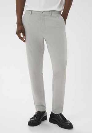 Homme portant un pantalon slim gris clair, une chemise blanche rentrée et des mocassins en cuir noir, debout les mains partiellement dans les poches.