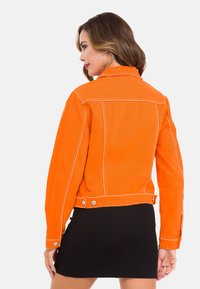 Orangene Jeansjacke mit weißen Nähten, einem Kragen und Druckknöpfen an der Taille, präsentiert an einem Modell, das einen schwarzen gerippten Rock trägt.