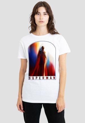 Young woman wearing a white T-shirt featuring a colorful, blurred, vertical Superman graphic and the text "SUPERMAN" at the bottom.