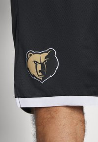 Shorts de sport noirs avec un tissu texturé et une bordure blanche. Un logo ours doré et noir est affiché de manière proéminente sur la cuisse gauche.