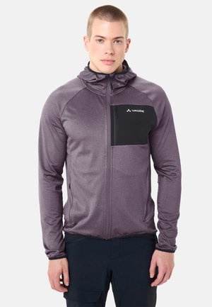 Junger Mann trägt einen lila Kapuzenpullover mit Reißverschluss und einer schwarzen Brusttasche mit der Aufschrift "Vaude", kombiniert mit dunkler Hose, stehend vor weißem Hintergrund.