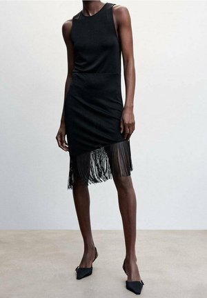 Femme portant une robe noire sans manches avec un ourlet à franges asymétrique et des escarpins à bout pointu noirs contre un arrière-plan uni.