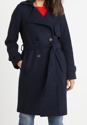 Frau, die einen marineblauen Wollmantel mit Gürtel und schwarzen Knöpfen über einem roten Oberteil und blauen Jeans trägt, die Hände in den Manteltaschen.