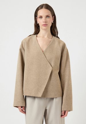 Frau mit langen braunen Haaren trägt eine beige Wickeljacke und hellgraue Hose, steht vor einem schlichten hellen Hintergrund.