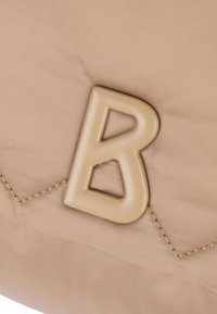 Beige tygväska med en framträdande beige bokstav "B" logotyp, quiltad struktur med synlig sömnad, slät yta.