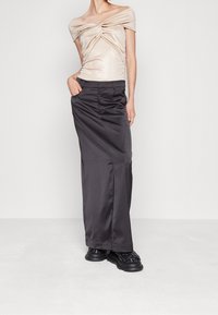 Kvinna klädd i en glittrig off-shoulder beige topp, svart högmidjad långkjol med fickor och kraftiga svarta vävda skor, stående mot en vit vägg.