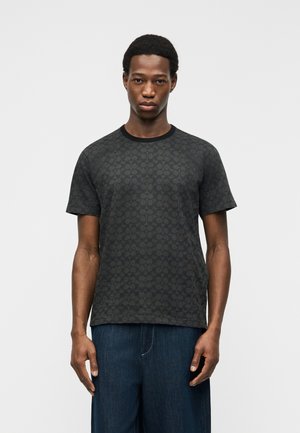 Young man wearing a black patterned crew neck t-shirt and dark blue jeans, standing against a plain light background.