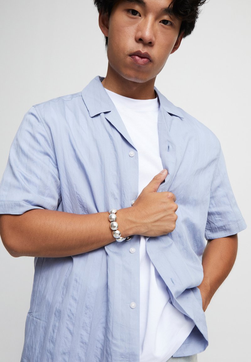 Chemise bleu clair à manches courtes avec une coupe décontractée, présentant une texture subtile et des détails de boutons, associée à un t-shirt blanc et un bracelet argenté.