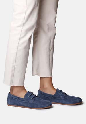 Fødder iført blå ruskindsmokkasiner kombineret med off-white cropped bukser, vist mod en hvid baggrund.