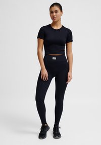 Schwarzes geripptes Kurzarm-Crop-Top, kombiniert mit hochgeschnittenen schwarzen Leggings. Beide Teile haben eine glatte Textur und ein kleines Markenetikett am Bündchen.