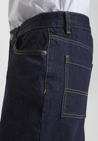 Tummansiniset farkut keltaisilla saumoilla, joissa on sivutasku, tavallinen vyötärönauha ja hienovarainen tekstuuri, nähtynä oikealta puolelta.