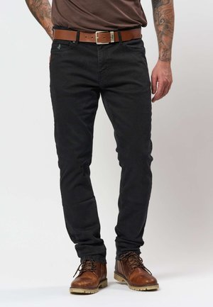 Férfi fekete slim-fit farmert, barna bőrtövet és barna fűzős csizmát visel, tetovált karokkal áll.