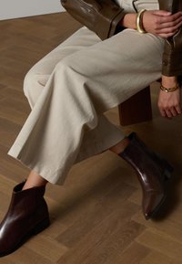 Brown leather ankle boot with pointed toe, low block heel, and side zipper. Worn with cream wide-leg pants on a wooden floor.