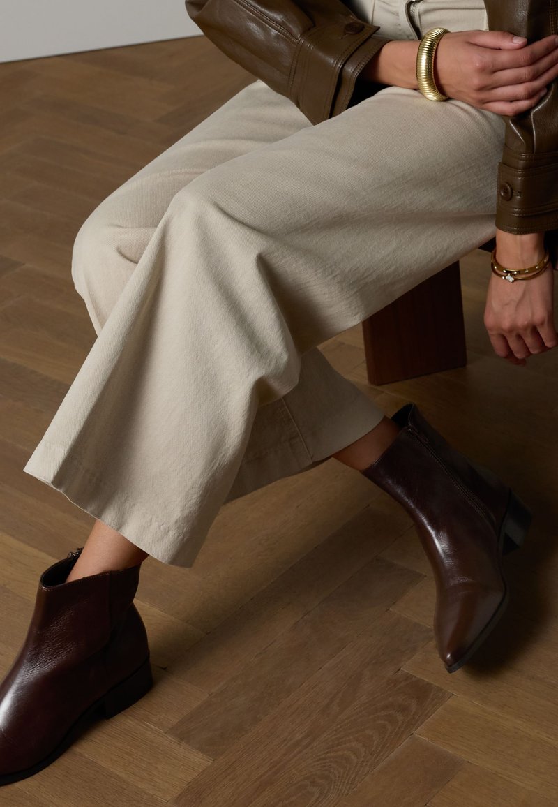 Brown leather ankle boot with pointed toe, low block heel, and side zipper. Worn with cream wide-leg pants on a wooden floor.