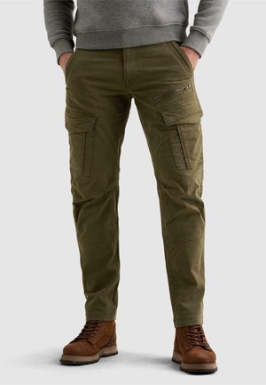 Man wearing olive green cargo pants with multiple pockets, grey sweatshirt, and brown lace-up boots standing with hands in pockets.