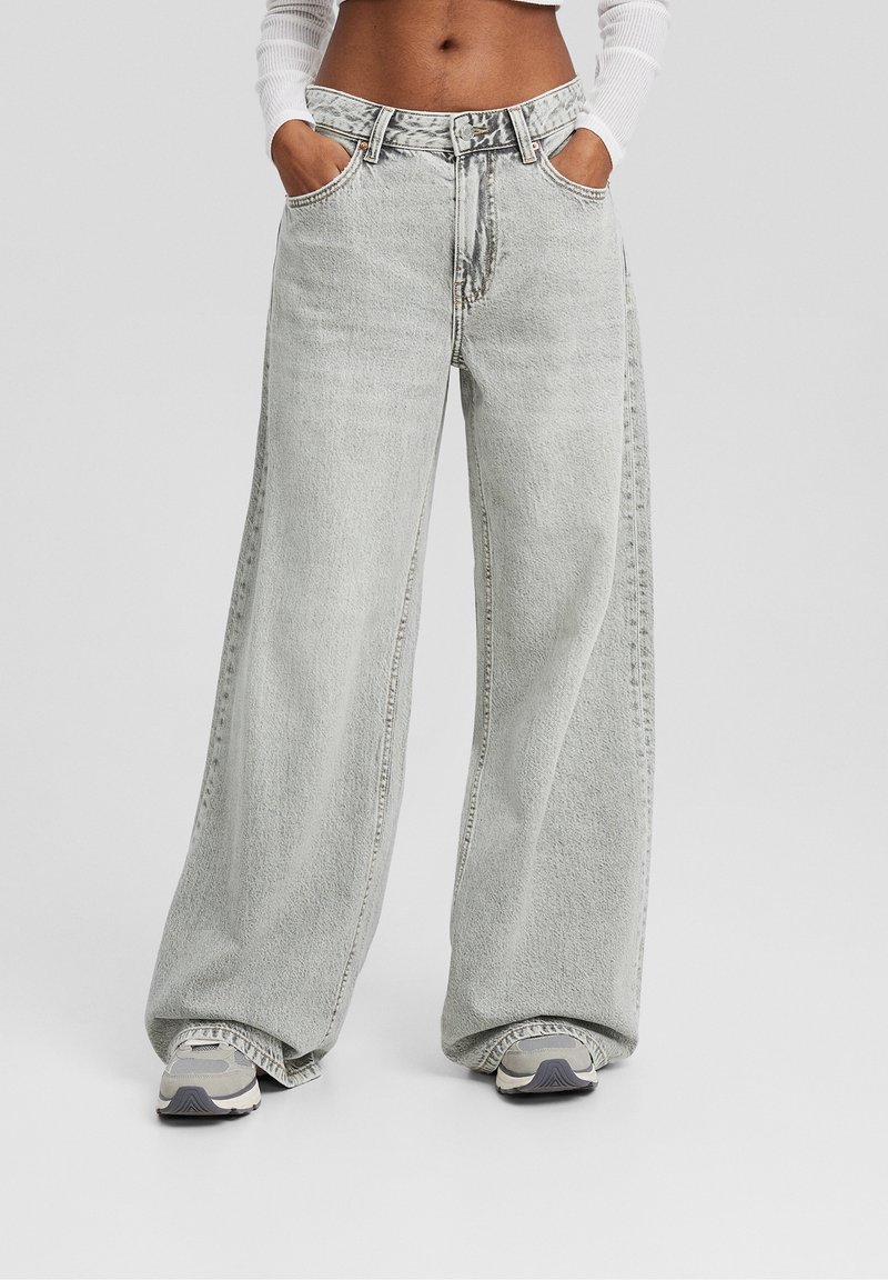 Personne portant un jean large gris clair et des baskets, les mains dans les poches, debout devant un fond clair uni.