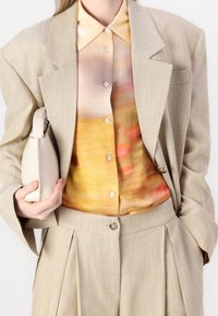Femme portant un blazer et un pantalon beige à plis, une blouse boutonnée multicolore, tenant un petit sac à main beige à ses côtés.