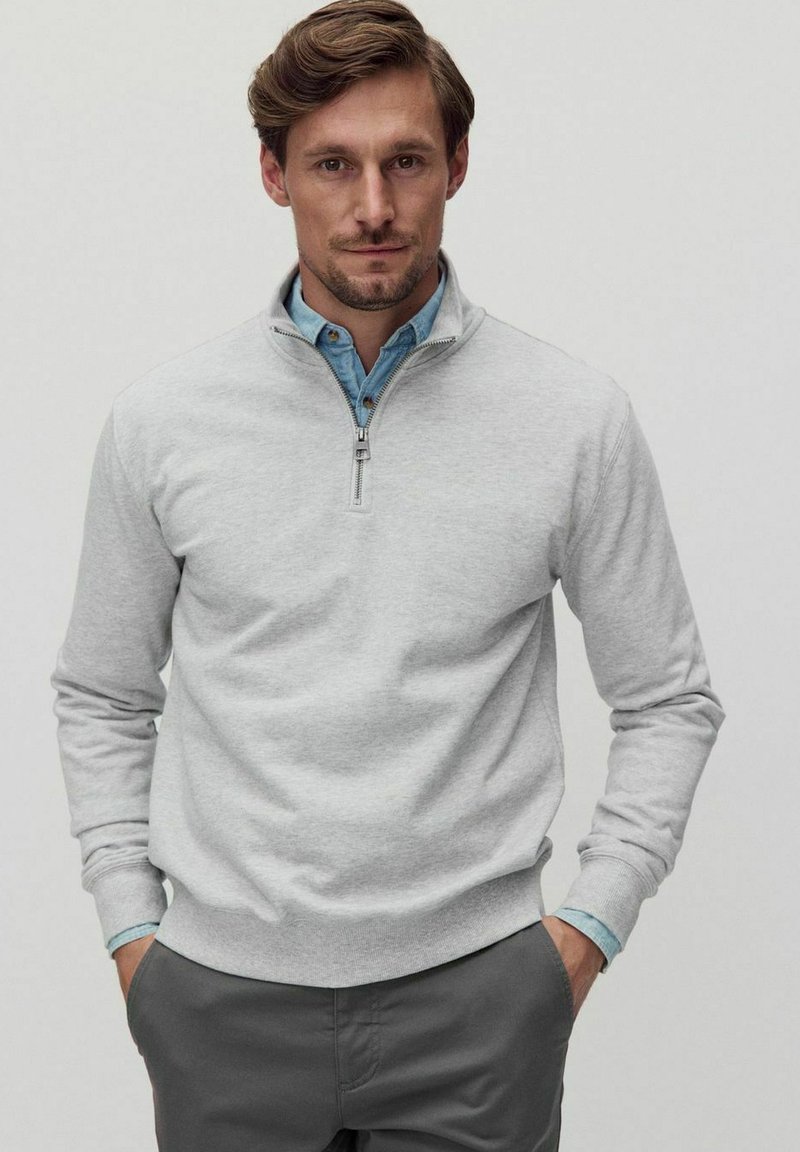Sweatshirt zippé gris clair avec une texture douce et des poignets côtelés, superposé sur une chemise bleue à col. Accompagné de pantalons gris.