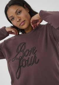 Burgundowa bluza z czarnym haftowanym napisem "Bonjour", okrągły dekolt i długie rękawy. Miękka tkanina o luźnym kroju.