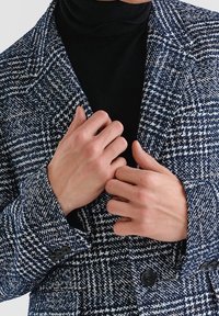 Navyblauw-witte houndstooth blazer met ingesneden revers, dubbele knopen en een gestructureerde stof. Draagt over een zwarte coltrui.