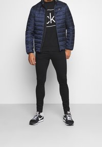 Chaqueta de nailon acolchada en color marino con cuello alto; sudadera negra con logo; jeans negros ajustados; zapatillas deportivas en blanco y negro.