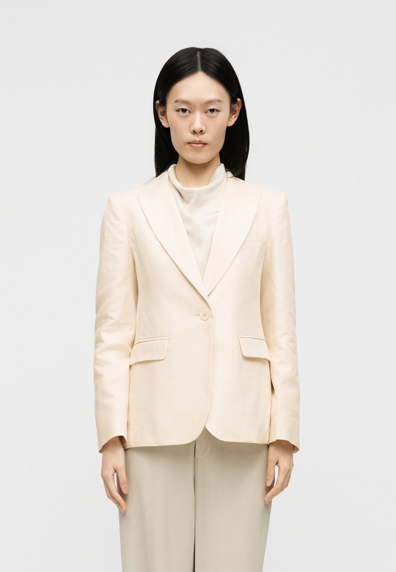 Femme aux cheveux noirs et raides, portant un blazer beige sur un top à col montant et un pantalon beige assorti, debout devant un fond blanc.