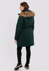Parka verde con cappuccio in pelliccia sintetica, vita elastica e tasche laterali. Indossata con leggins neri e sneakers. Tessuto liscio, taglio a lunghezza media.