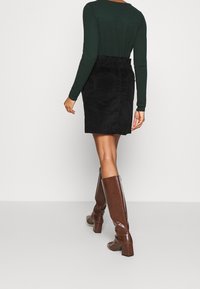 Svart randig manchesterkjol med fickor, kombinerad med en texturerad mörkgrön topp och glansiga bruna knähöga stövlar med klack.