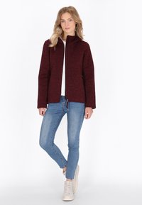 Maroon jacket with a high collar, zipper front, and long sleeves. Worn over a white top and paired with blue jeans and light sneakers.