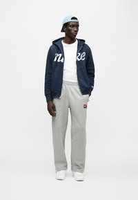 Νέα μπλε φούτερ με φερμουάρ και λευκή επιγραφή "Nike", γκρι φόρμα με λογότυπο, λευκό μπλουζάκι και γαλάζιο καπέλο, στέκεται σε ουδέτερο φόντο.