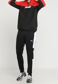 Puma zwarte en rode windbreaker met een voorzak, gecombineerd met zwarte broek met witte zijstrepen en ritsen bij de enkels.