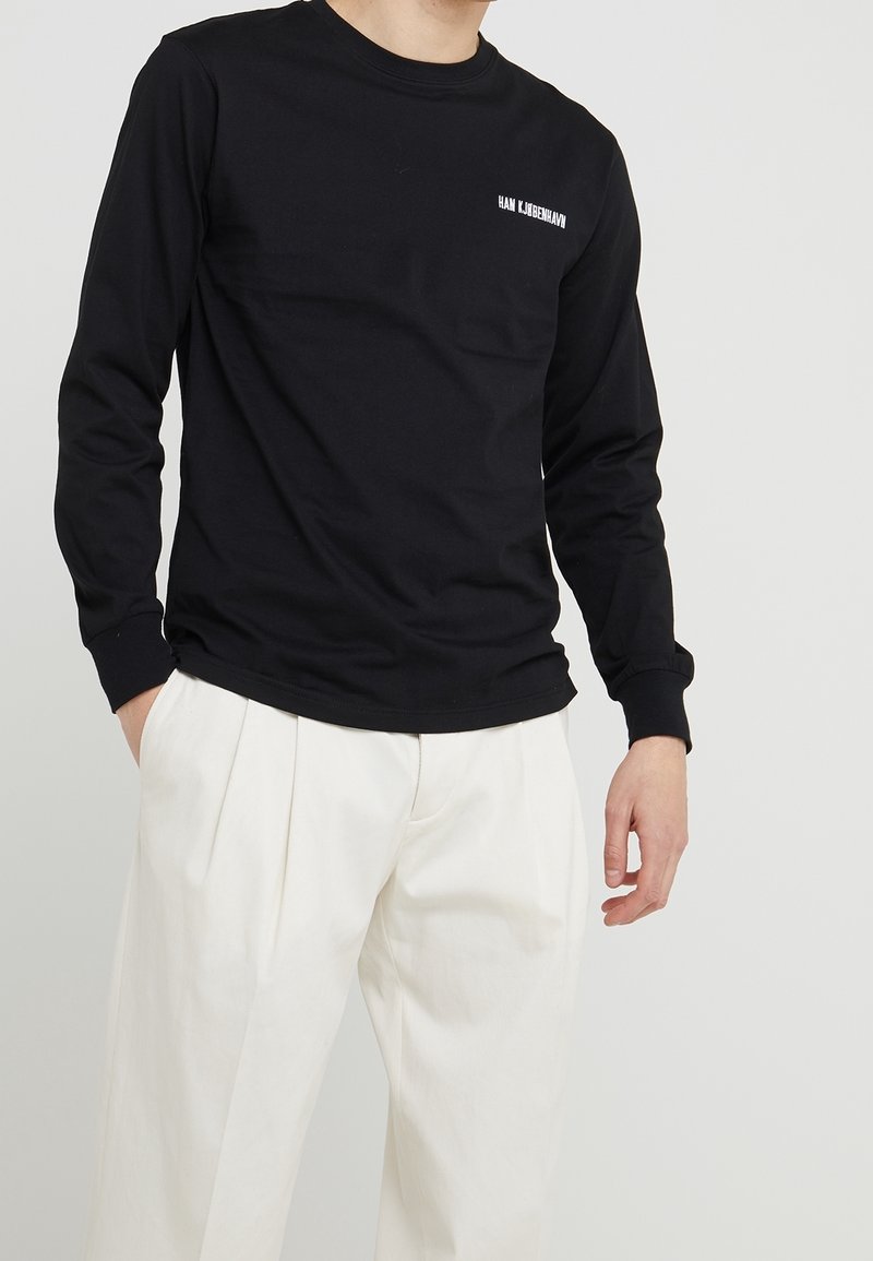 Svart långärmad t-shirt med rund halsringning och vitt tryckt varumärke på vänster bröst, kombinerad med beige veckade byxor.