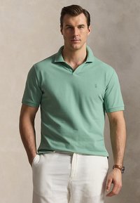 Mann mit kurzen braunen Haaren, der ein hellgrünes Poloshirt und weiße Hosen trägt, vor einem schlichten beigen Hintergrund stehend.