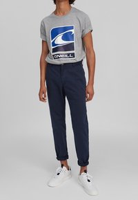 Osoba nosząca szary t-shirt z logo O'Neill, granatowe spodnie z podwiniętymi nogawkami i białe trampki na jednolitym szarym tle.