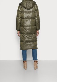 Abrigo largo acolchado de color verde oliva con capucha, con un acabado brillante, diseño acolchado y puños elásticos, combinado con vaqueros azules y botines beige.