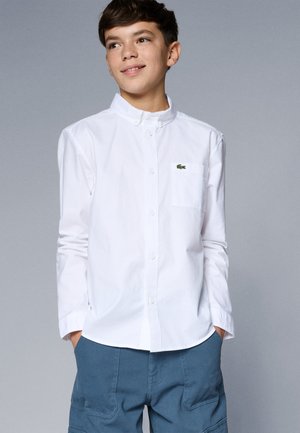 Camicia bianca con colletto, dotata di una tasca sul petto e un piccolo logo verde. Il tessuto sembra morbido e leggero.