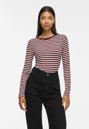Junge Frau mit langen dunklen Haaren, die ein bordeauxrot-weiß gestreiftes Langarmshirt und hochtaillierte schwarze Hosen trägt, steht vor weißem Hintergrund.