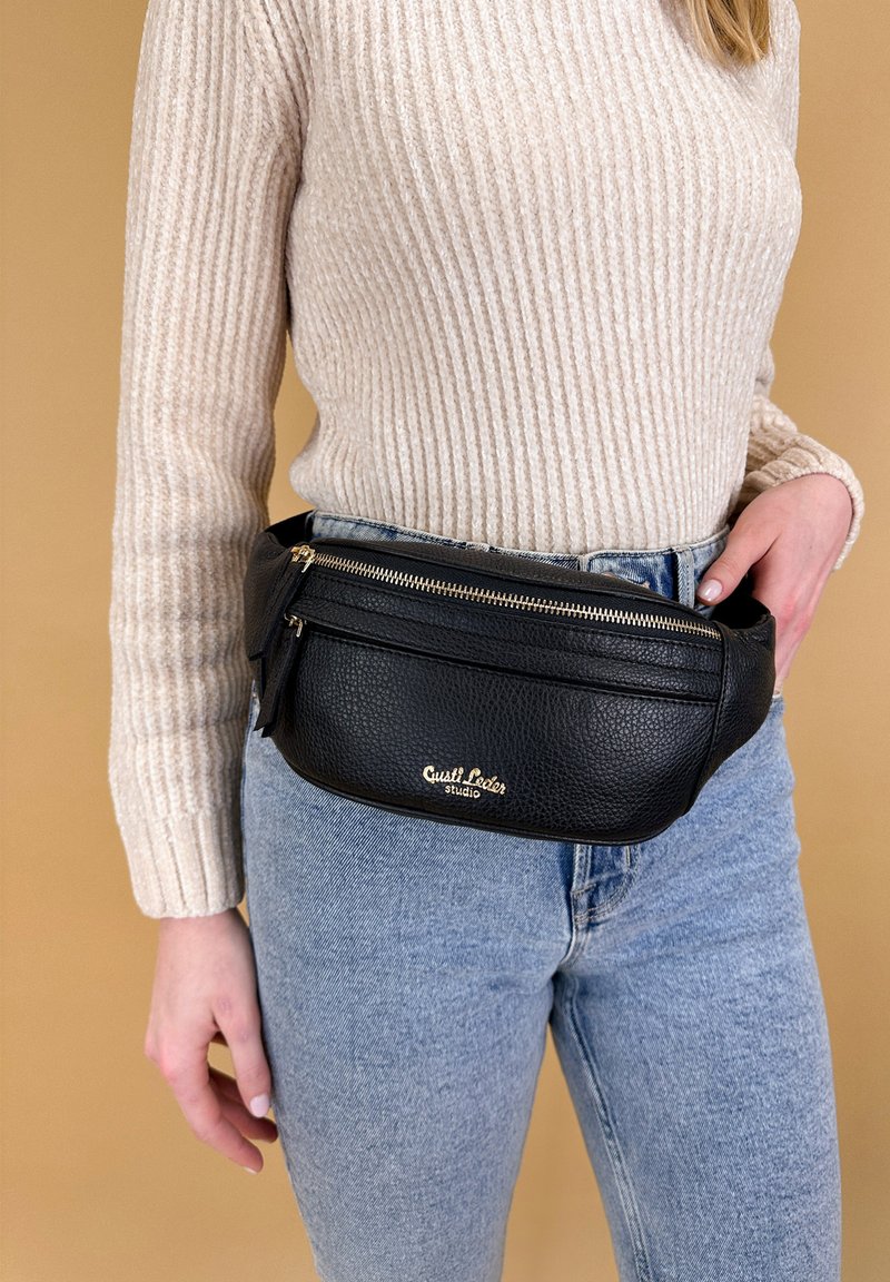 Persoon in een beige gebreide trui en blauwe jeans met een zwarte leren heuptas met gouden ritsen en logo aan de voorkant.
