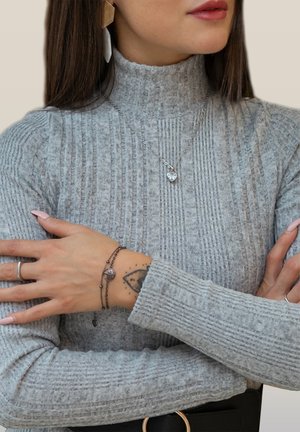 Grau gestreifter Rollkragenpullover mit langen Ärmeln, mehrlagige Silberkette mit einem Herzanhänger und ein Armband mit dekorativen Elementen.