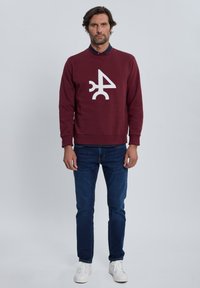 Sudadera color granate con un diseño gráfico blanco, combinada con vaqueros azul oscuro y zapatillas blancas. Tejido de textura suave, cuello redondo.