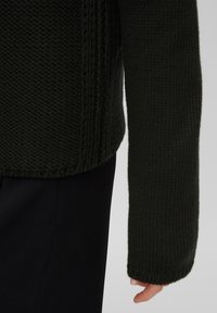 Mörkgrön stickad tröja med ett texturerat mönster, med långa ärmar och ribbade muddkanter, kombinerad med svart tyg under.