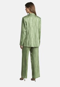 Groen satijnen pak met een textuurafwerking, bestaande uit een lange blazer en wijde broek, beide met een subtiel patroon en een op maat gemaakte uitstraling.