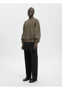 Oļīvzaļš džemperis ar nolaistām pleciem un ribveida aprocēm, kopā ar melnām plaša piegriezuma biksēm un spīdīgām melnām kurpēm.