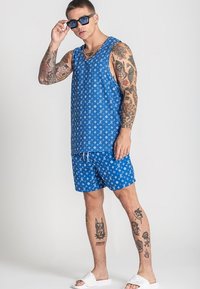 Modrá tielko a šortky so opakujúcim sa bielym vzorom; tkanina sa zdá byť ľahká. Doplnky zahŕňajú tmavé slnečné okuliare a biele šľapky.